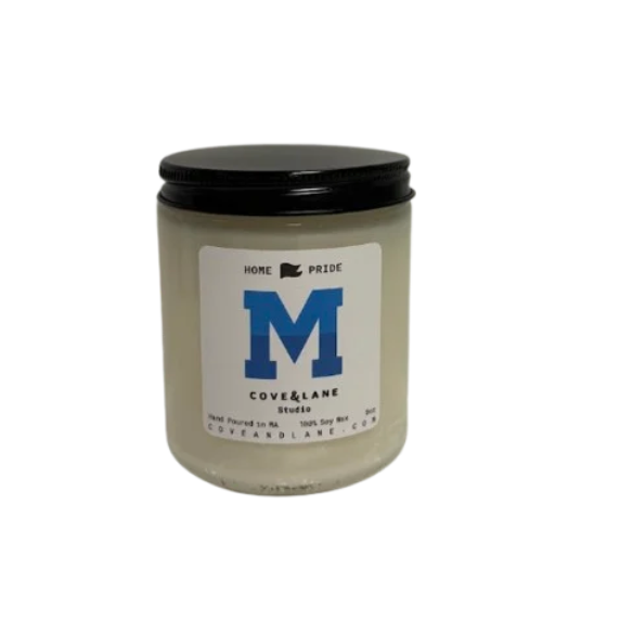 A jar candle with a black lid and a white label featuring a large blue letter M and the text 'HOME PRIDE COVE & LANE Studio'. Available at Be Chamred