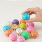 Colorful dumpling-shaped toys with a hand picking one up on a white background. Available at Be Charmed