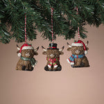 Three decorative Highland cow ornaments with festive hats and scarves hanging from a Christmas tree. Available at Be Charmed