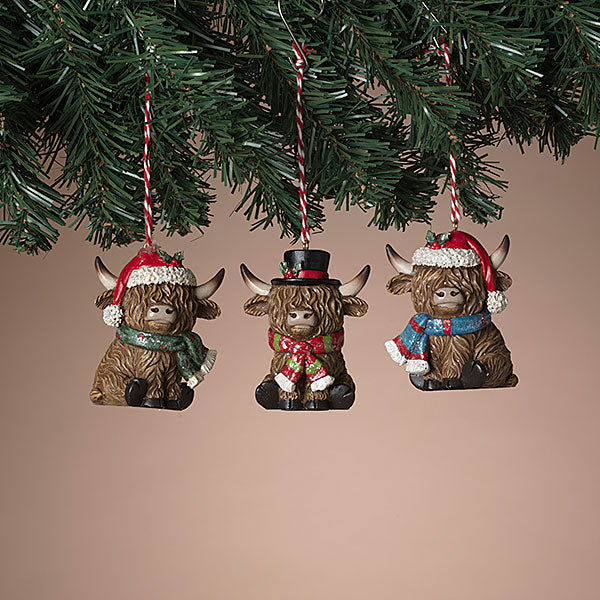 Three decorative Highland cow ornaments with festive hats and scarves hanging from a Christmas tree. Available at Be Charmed