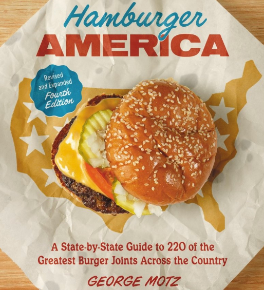 Book cover of 'Hamburger America' with a burger illustration on a wooden surface. Available at Be Charmed