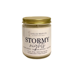 A jar candle from Lola & Bean Co. named 'Stormy nights', featuring a cream-colored label with black text and a metallic gold screw-on lid, set against a plain white background. Available at Be Charmed