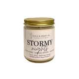A jar candle from Lola & Bean Co. named 'Stormy nights', featuring a cream-colored label with black text and a metallic gold screw-on lid, set against a plain white background. Available at Be Charmed