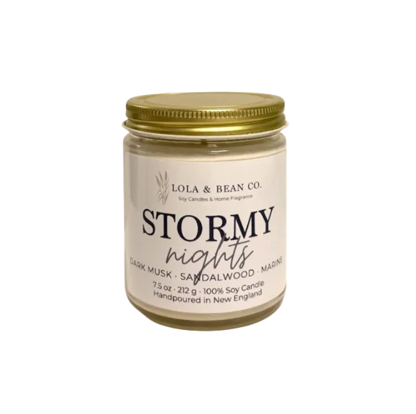 A jar candle from Lola & Bean Co. named 'Stormy nights', featuring a cream-colored label with black text and a metallic gold screw-on lid, set against a plain white background. Available at Be Charmed