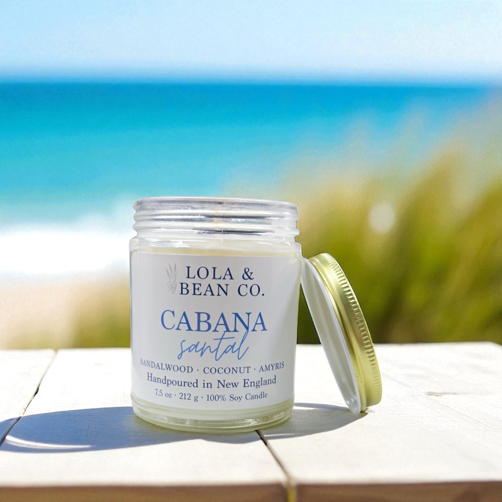 Candle jar labeled 'Lola & Bean Co. Cabana Santal' on a wooden surface with a blurred beach background. Available at Be Charmed