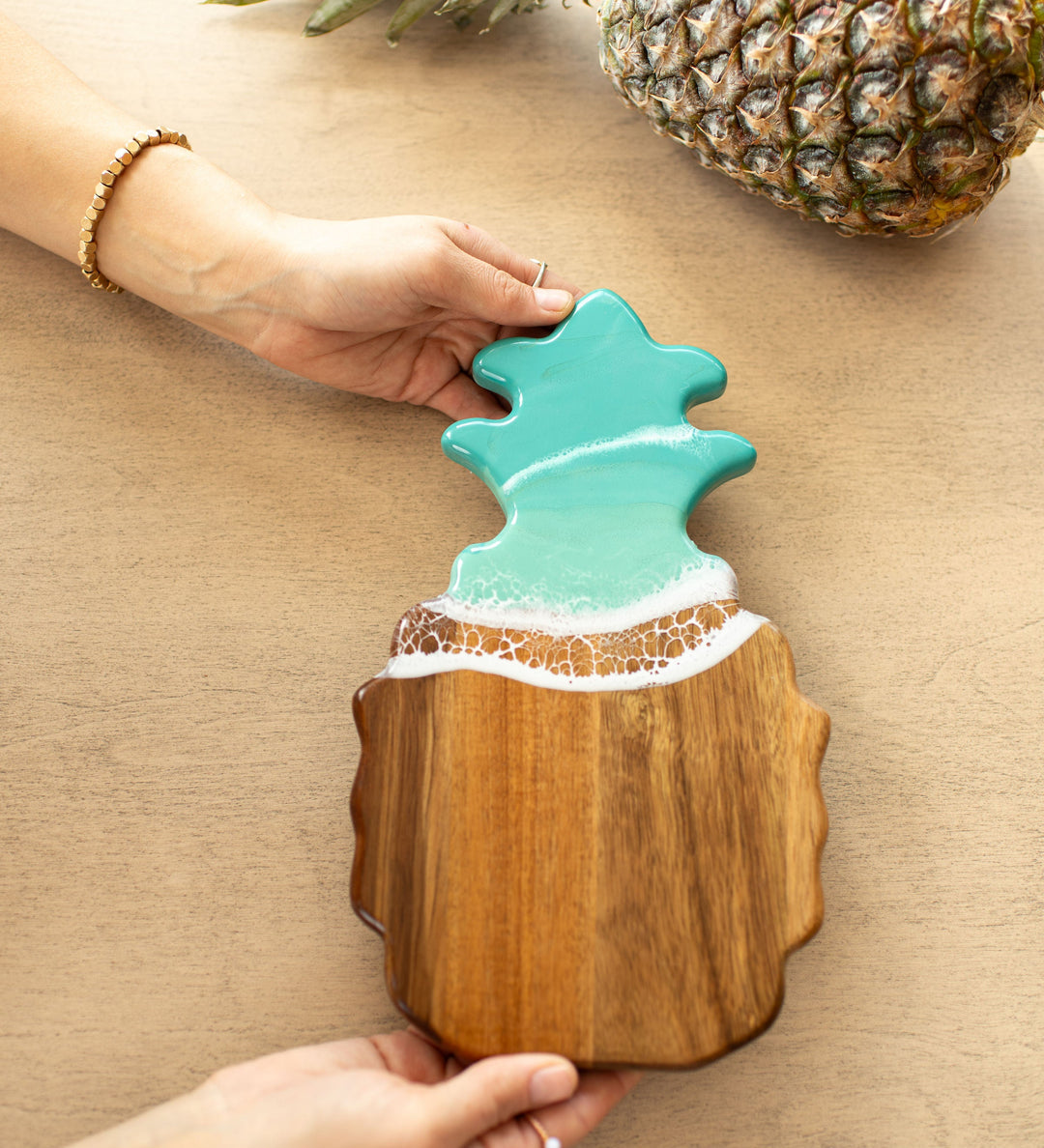 Wooden caribbean blue pineapple-shaped cutting board with a decorative top on a beige surfaceb. Available at Be Charmed