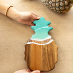 Wooden caribbean blue pineapple-shaped cutting board with a decorative top on a beige surfaceb. Available at Be Charmed