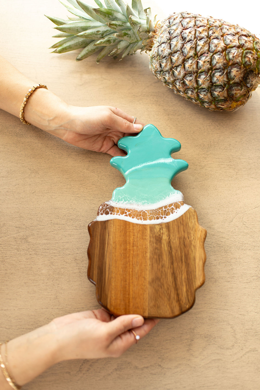 Wooden caribbean blue pineapple-shaped cutting board with a decorative top on a beige surfaceb. Available at Be Charmed