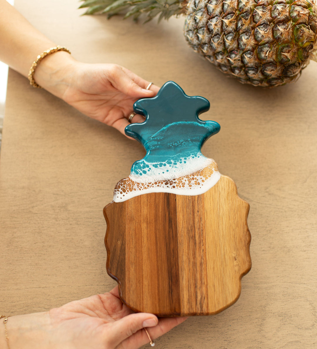 Wooden ocean vibes pineapple-shaped cutting board with a blue resin top held by a person on a beige surface. Availabe at Be Charmed