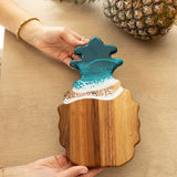 Wooden ocean vibes pineapple-shaped cutting board with a blue resin top held by a person on a beige surface. Availabe at Be Charmed