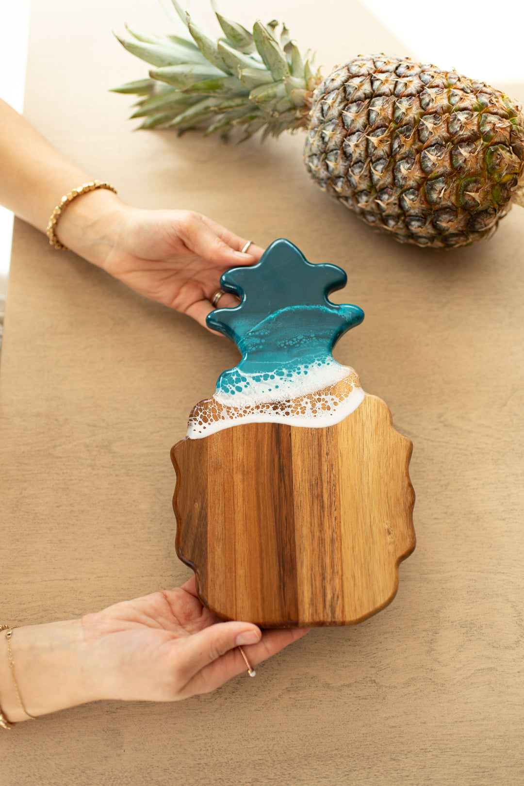 Wooden ocean vibes pineapple-shaped cutting board with a blue resin top held by a person on a beige surface. Availabe at Be Charmed