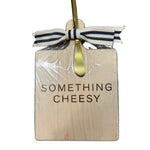 Wooden cutting board with 'Something Cheesy' text, gold fork, and ribbon on a wooden surface. Available at Be Charmed