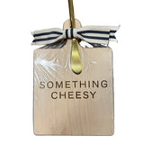 Wooden cutting board with 'Something Cheesy' text, gold fork, and ribbon on a wooden surface. Available at Be Charmed