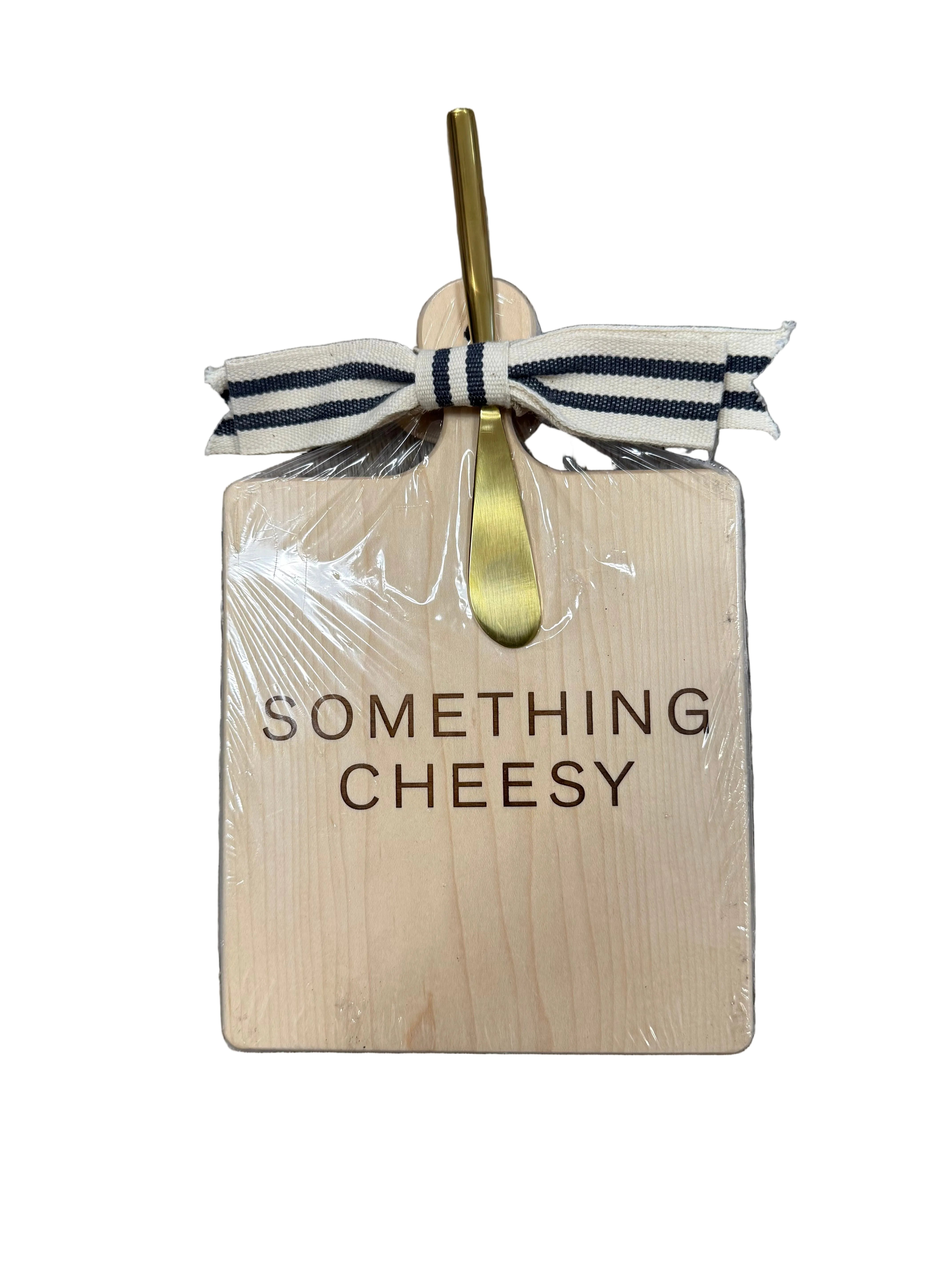Wooden cutting board with 'Something Cheesy' text, gold fork, and ribbon on a wooden surface. Available at Be Charmed
