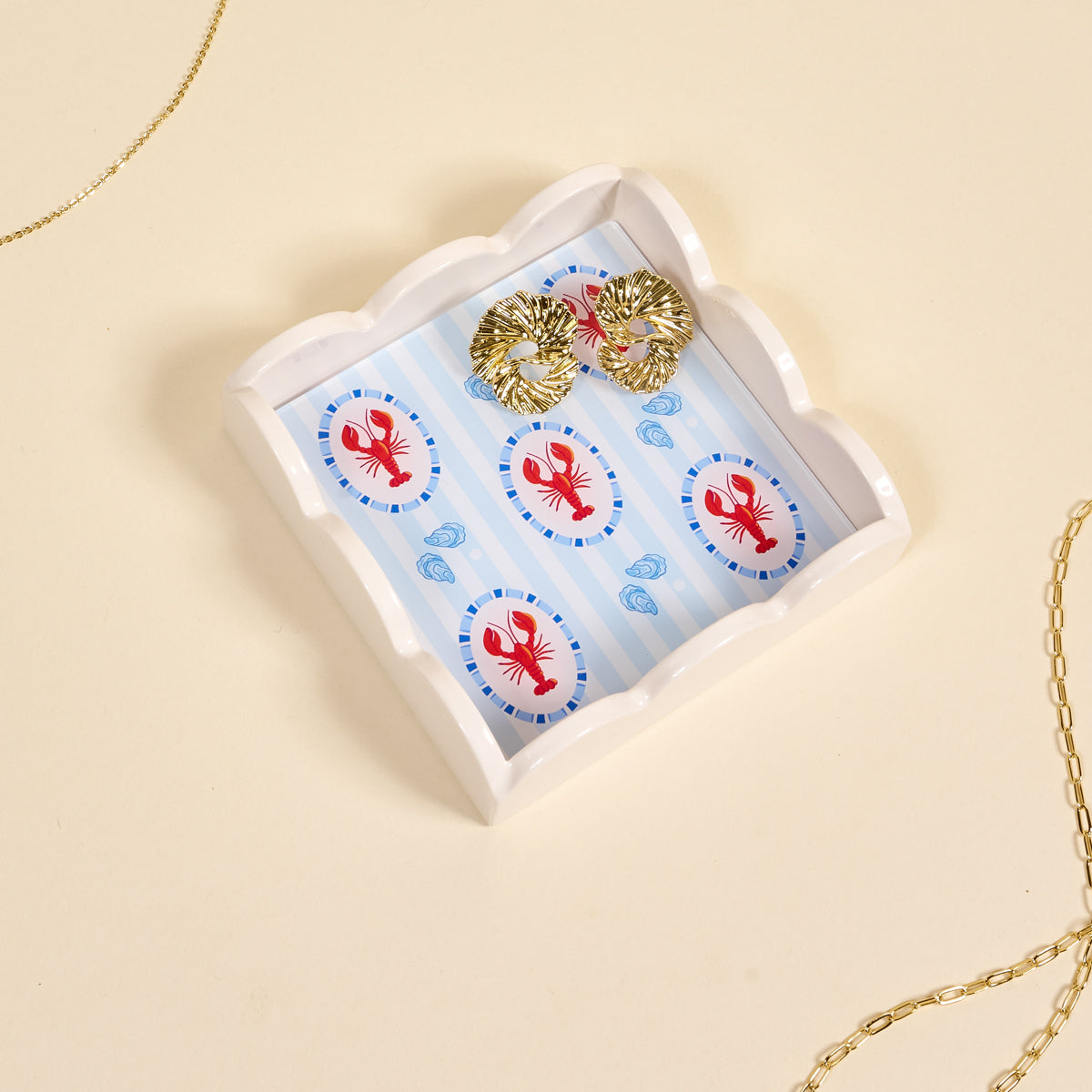 Gold earrings on a decorative tray with lobster design on a beige background. Available at Be Charmed