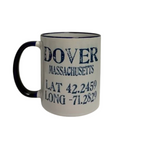 A white ceramic coffee mug with a navy handle and navy rim, printed with the text 'DOVER MASSACHUSETTS' and coordinates in dark blue lettering. Available at Be Charmed