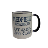 A white ceramic mug with a navy handle and rim, printed with the text 'MEDFIELD MASSACHUSETTS' and coordinates 'LAT 42.1876' and 'LONG -71. Available at Be Charmed
