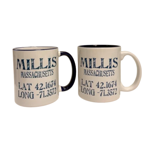Two ceramic mugs on a white background, one showing the black handle and the other showing the white handle. Both mugs are cream-colored with navy interiors and printed with the text 'MILLIS MASSACHUSETTS' and coordinates 'LAT 42. Available at Be Charmed