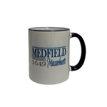 A white ceramic coffee mug with a navy handle and rim, featuring the text 'MEDFIELD ESTABLISHED 1649 Massachusetts' printed in navy blue and gray on the side. Available at Be Charmed