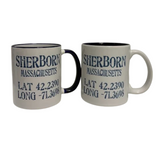 Two white ceramic mugs with navy interiors and handles, both printed with the text 'SHERBON MASSACHUSETTS LAT 42.2390 LONG -71. Available at Be Charmed