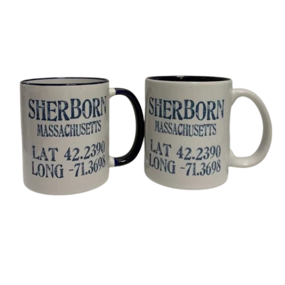 Two white ceramic mugs with navy interiors and handles, both printed with the text 'SHERBON MASSACHUSETTS LAT 42.2390 LONG -71. Available at Be Charmed