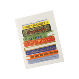 A birthday greeting card featuring a stack of colorful illustrated books with text including 'HERE'S TO ANOTHER Card Charlotte', 'HAPPY BIRTHDAY', and a quote from Mary Anne Patterson, shown on a light gray background. Available at Be Charmed