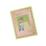 A greeting card with a vintage illustration of a girl in a blue dress standing beside a tiered birthday cake, framed by a pink lace-like border and a pale yellow outer border with floral details. The card is photographed at an angle on a clean white background. Available at Be Charmed