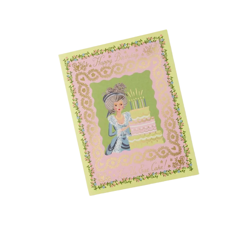 A greeting card with a vintage illustration of a girl in a blue dress standing beside a tiered birthday cake, framed by a pink lace-like border and a pale yellow outer border with floral details. The card is photographed at an angle on a clean white background. Available at Be Charmed