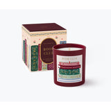 A Rifle Paper Co red glass jar candle labeled 'Book Club' stands in front of its matching illustrated box on a clean white background. The box features vintage-style book spines. Available at Be Charmed