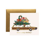 A Rifle Paper Co holiday greeting card with a gold envelope, featuring an illustration of a wood-paneled car carrying a Christmas tree and wrapped gifts, with the text 'Have a Jolly Holiday!' on a white background. Available at Be Charmed
