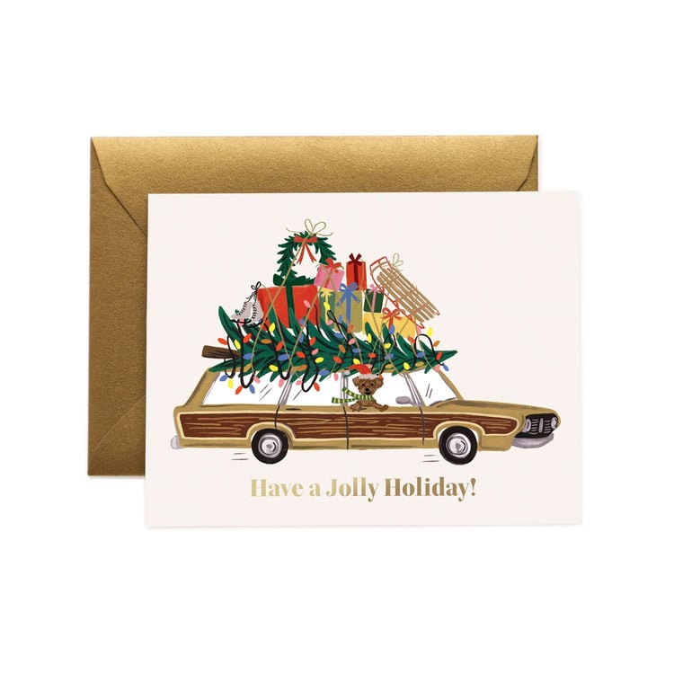 A Rifle Paper Co holiday greeting card with a gold envelope, featuring an illustration of a wood-paneled car carrying a Christmas tree and wrapped gifts, with the text 'Have a Jolly Holiday!' on a white background. Available at Be Charmed
