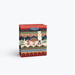 A Rifle Paper Co  gift bag with a colorful winter holiday pattern featuring a snowman, a cabin, and festive Fair Isle-style borders, shown on a light gray background. Available at Be Charmed
