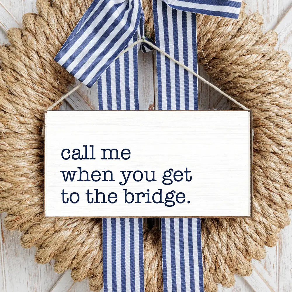 A rustic wooden sign with the phrase 'call me when you get to the bridge.' surrounded by decorative twine and blue and white striped ribbons. Available at Be Charmed