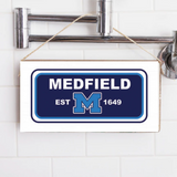 A rectangular wooden sign with 'MEDFIELD EST. 1649' printed in blue and white, hanging on a shower wall. Available at Be Charmed