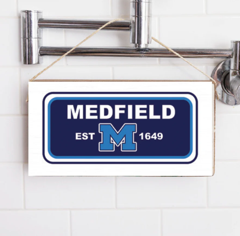 A rectangular wooden sign with 'MEDFIELD EST. 1649' printed in blue and white, hanging on a shower wall. Available at Be Charmed