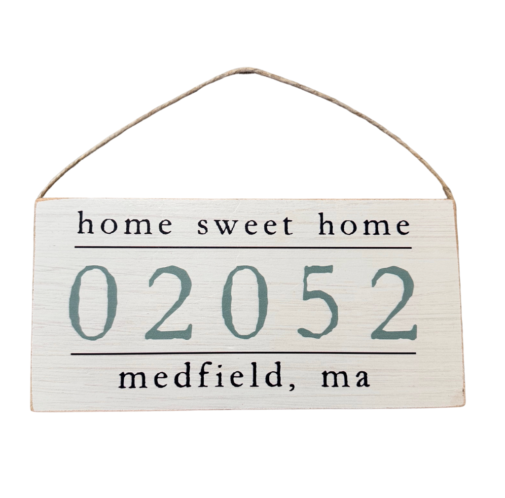 A wooden hanging sign with 'Home Sweet Home' text and a zip code '02052 Medfield, MA' below it. Available at Be Charmed