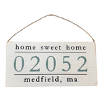A wooden hanging sign with 'Home Sweet Home' text and a zip code '02052 Medfield, MA' below it. Available at Be Charmed