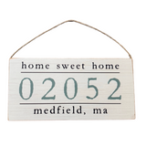 A wooden hanging sign with 'Home Sweet Home' text and a zip code '02052 Medfield, MA' below it. Available at Be Charmed