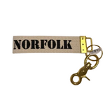 A canvas keychain strap with the word 'NORFOLK' printed in bold navy letters, attached to a gold-tone metal clasp and key rings on a white background. Available at Be Charmed