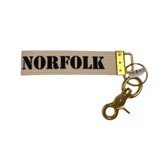 A canvas keychain strap with the word 'NORFOLK' printed in bold navy letters, attached to a gold-tone metal clasp and key rings on a white background. Available at Be Charmed