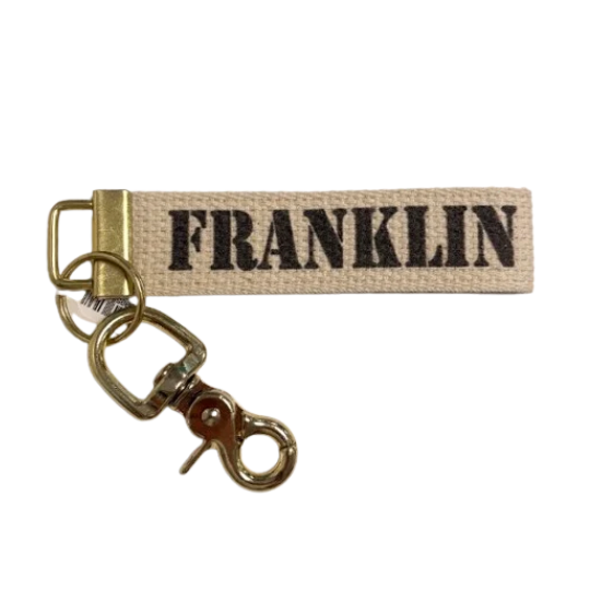 A canvas keychain or bag tag with the word 'FRANKLIN' printed in black capital letters on a beige woven fabric strap, attached to a gold-tone metal swivel clasp on a white background. Available at Be Charmed