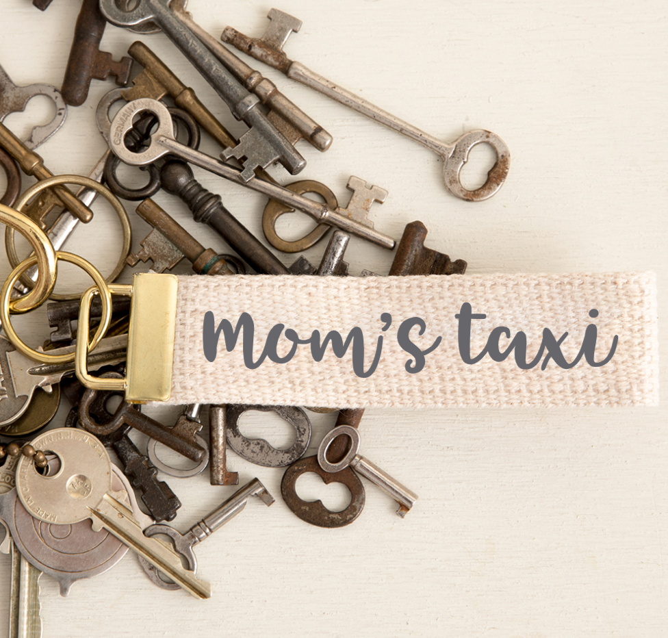 A gold-toned Rustic Marlin metal keychain clip with the text 'Mom's Taxi' printed on a beige cotton webbing strap, surrounded by various keys. Available at Be Charmed