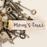 A gold-toned Rustic Marlin metal keychain clip with the text 'Mom's Taxi' printed on a beige cotton webbing strap, surrounded by various keys. Available at Be Charmed
