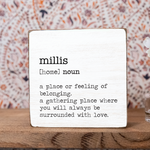 A decorative wooden block with the definition of 'millis' printed on it, placed on a wooden surface with a patterned background. Available at Be Charmed