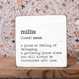 A decorative wooden block with the definition of 'millis' printed on it, placed on a wooden surface with a patterned background. Available at Be Charmed