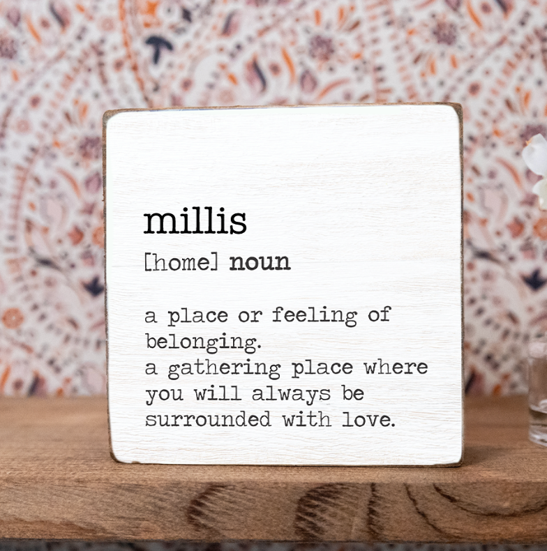 A decorative wooden block with the definition of 'millis' printed on it, placed on a wooden surface with a patterned background. Available at Be Charmed