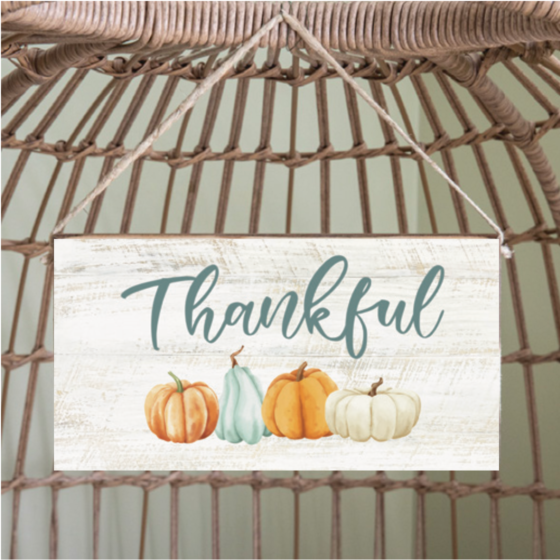 A rustic marlin style twine hanging sign with the word 'Thankful' written on it, accompanied by images of pumpkins in various colors. The sign is hanging from a twine and is placed against a woven background. Available at Be Charmed
