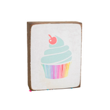 A decorative wooden block with a cupcake design, featuring a multicolored icing and a cherry on top, placed on a surface with colorful sprinkles scattered around. Available at Be Charmed