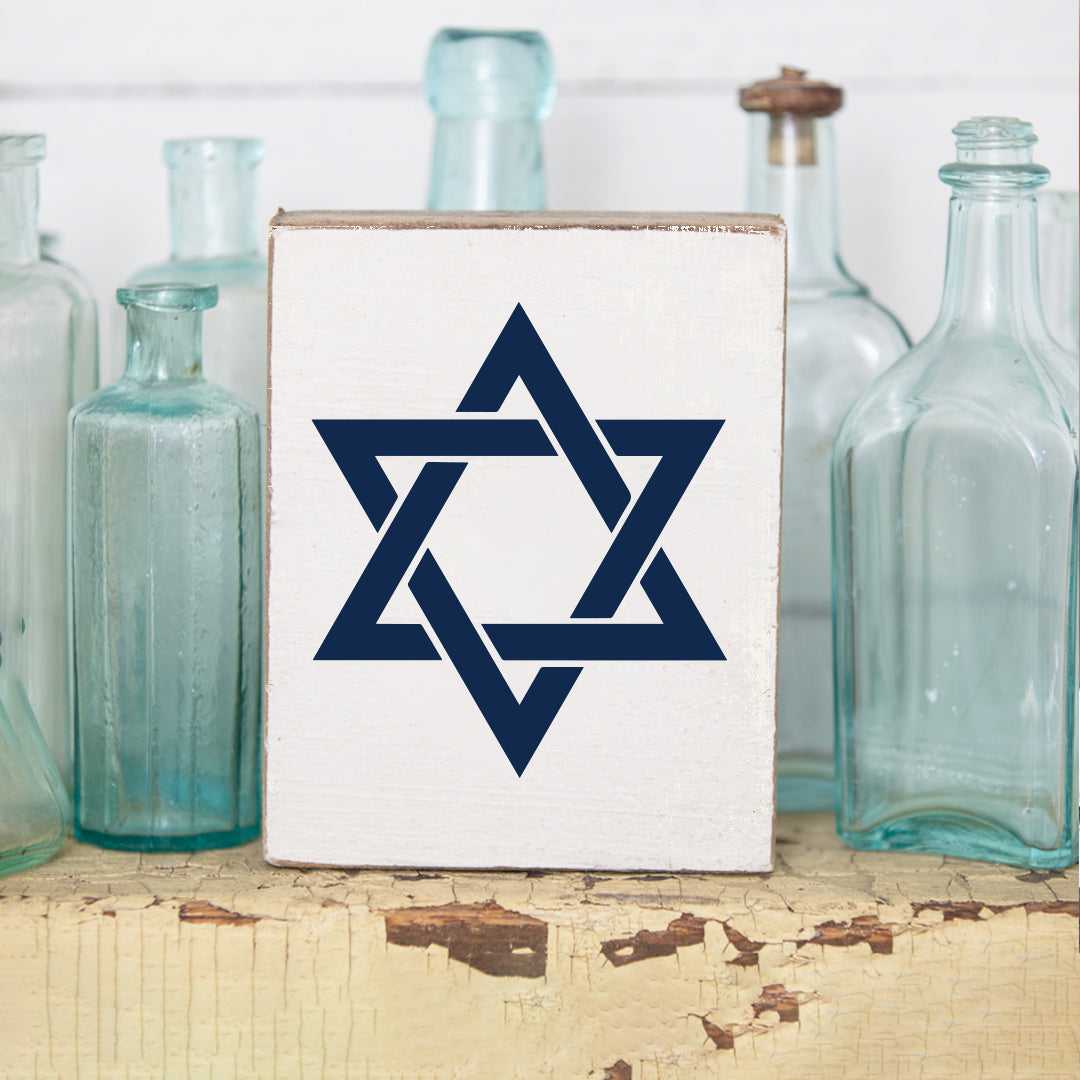Decorative wooden block with the Star of David design, placed on a rustic wooden surface with glass bottles in the background. Available at Be Charmed