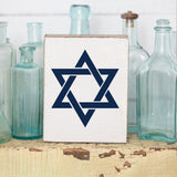 Decorative wooden block with the Star of David design, placed on a rustic wooden surface with glass bottles in the background. Available at Be Charmed